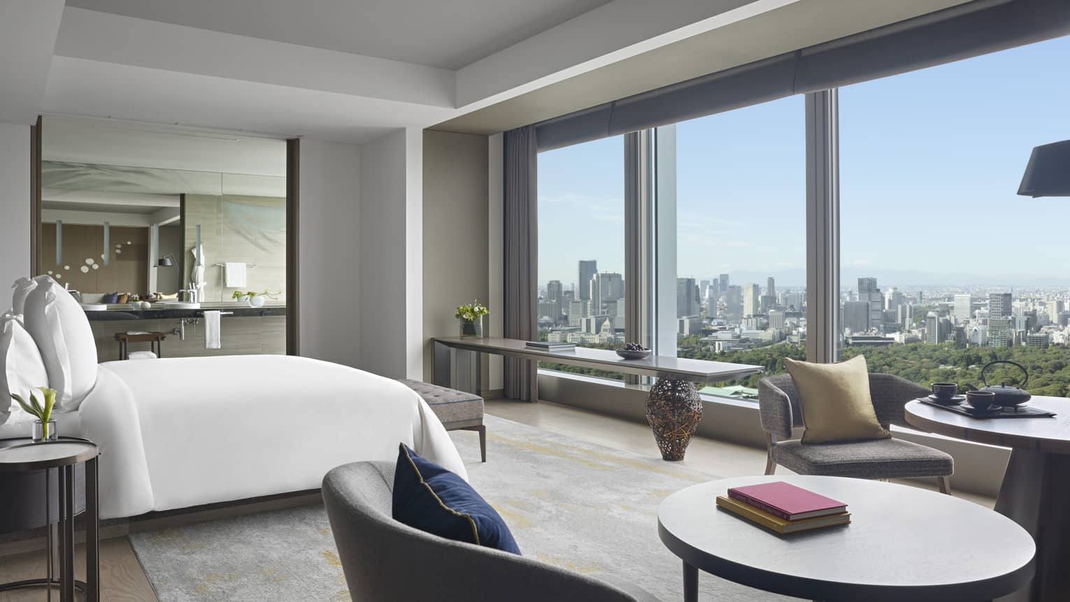 Four Seasons hotel suite with king bed, seating area with round tables and armchairs, side table, large mirror and windows with city views