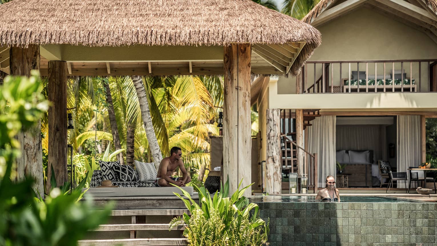 A smiling couple enjoy a pool and cabana outside of a villa on a sunny day. The villa is surrounded by trees and greenery.
