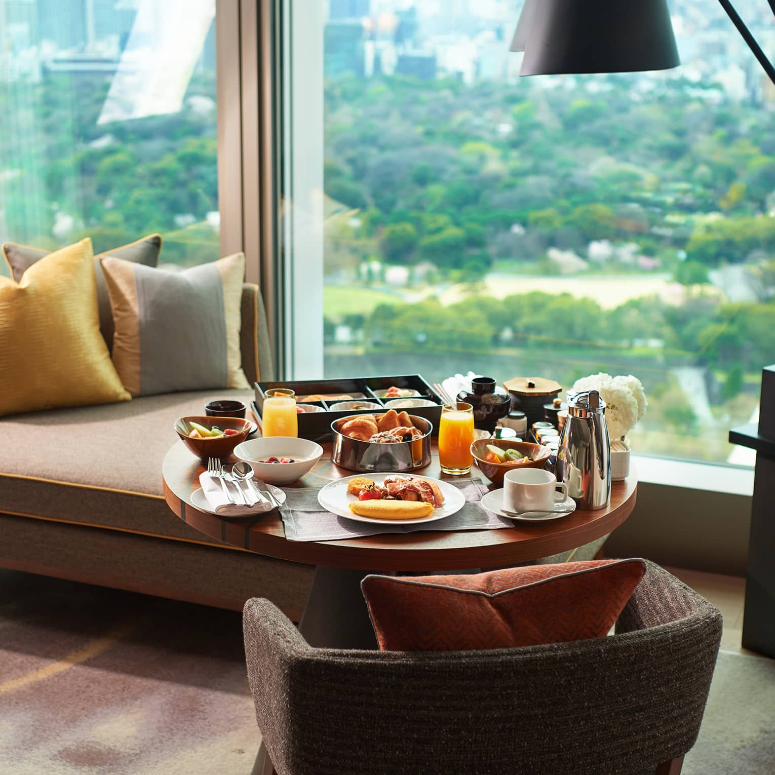 A breakfast tray of pastries, fresh fruit, juice and coffee on a guest room lounge chair at a window overlooking lush gardens.