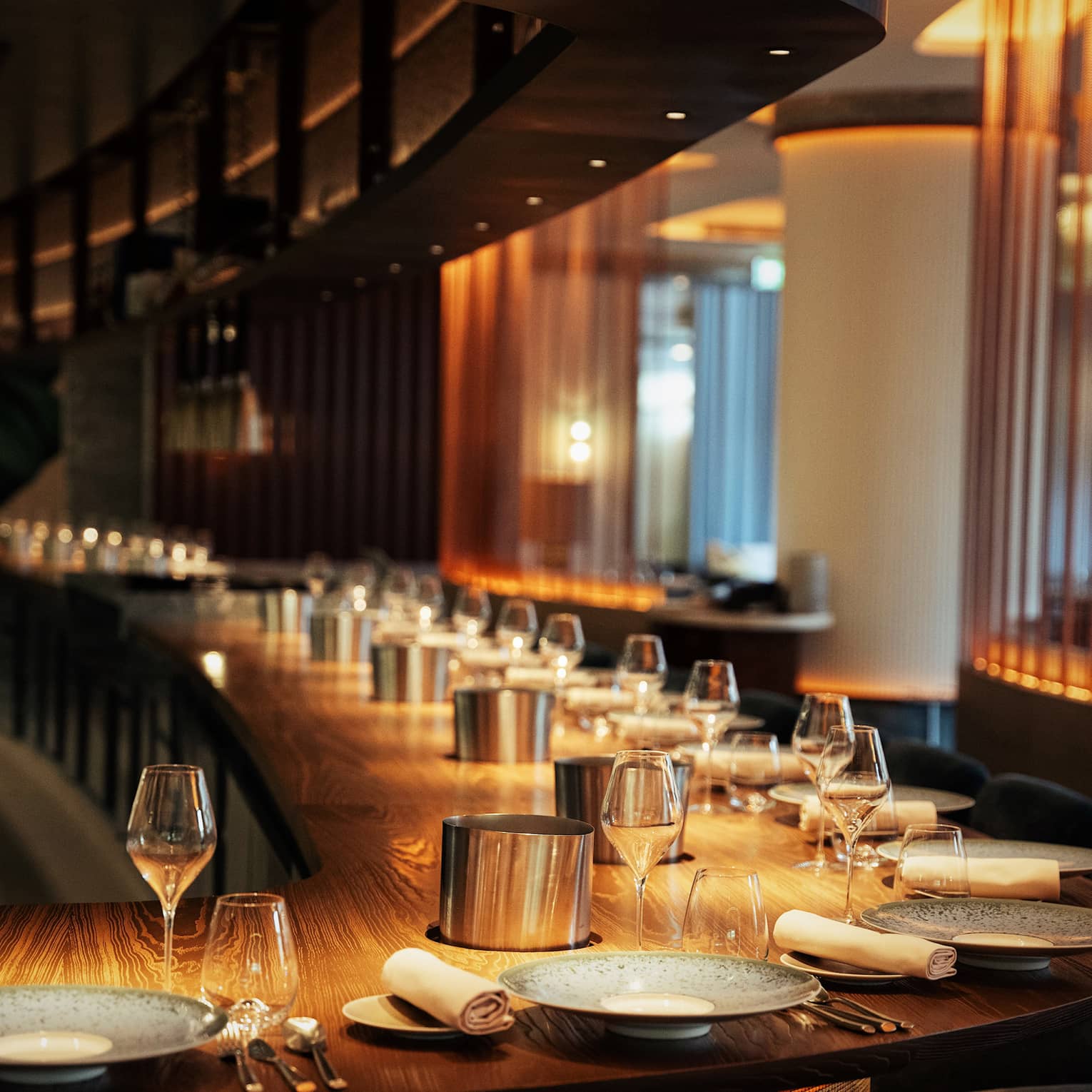 A long, curved solid wood bar with a dozen place settings in warm, golden lighting.