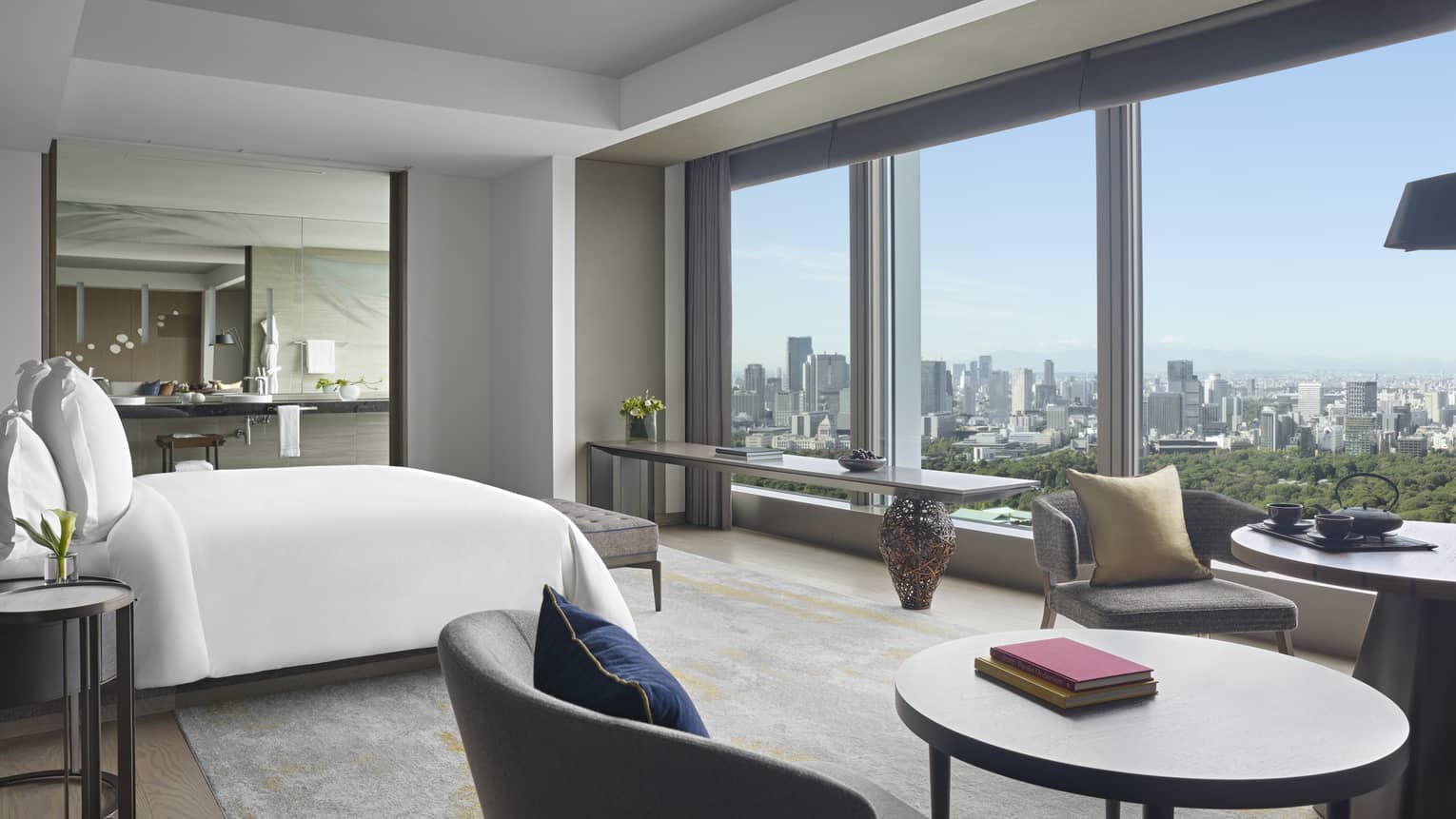 Four Seasons hotel suite with king bed, seating area with round tables and armchairs, side table, large mirror and windows with city views