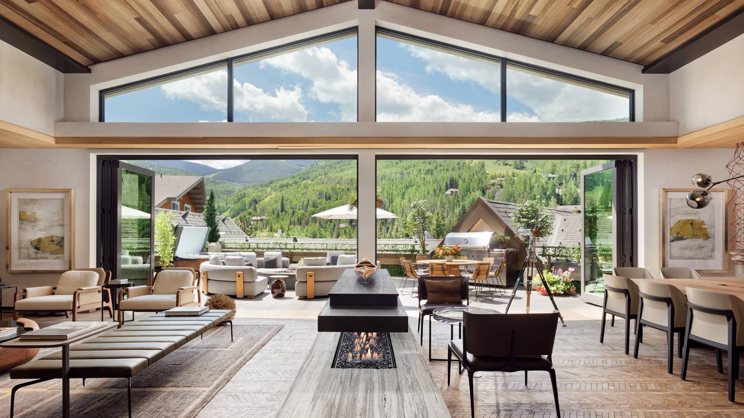 Luxurious mountain villa interior with an open-plan living area, modern fireplace, and expansive glass doors leading to an outdoor terrace with panoramic forest views.