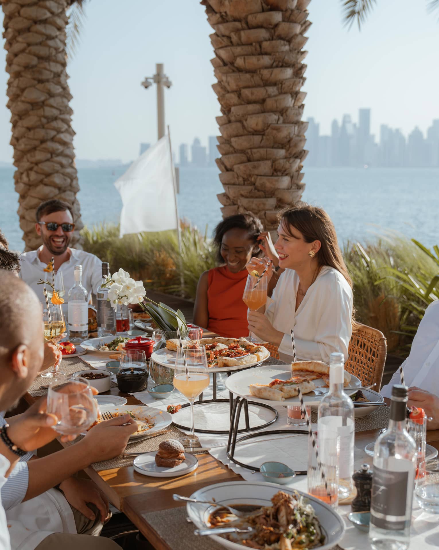 Group sits around long outdoor dining table eating brunch with the ocean and a city skyline in the background