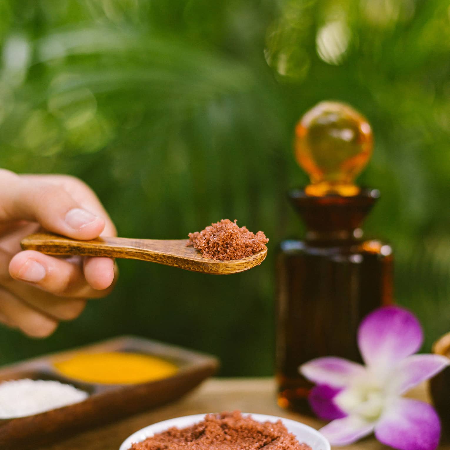 Hand holds small wood spoon with salt scrub over bowl on wood spa table with flower, glass bottle