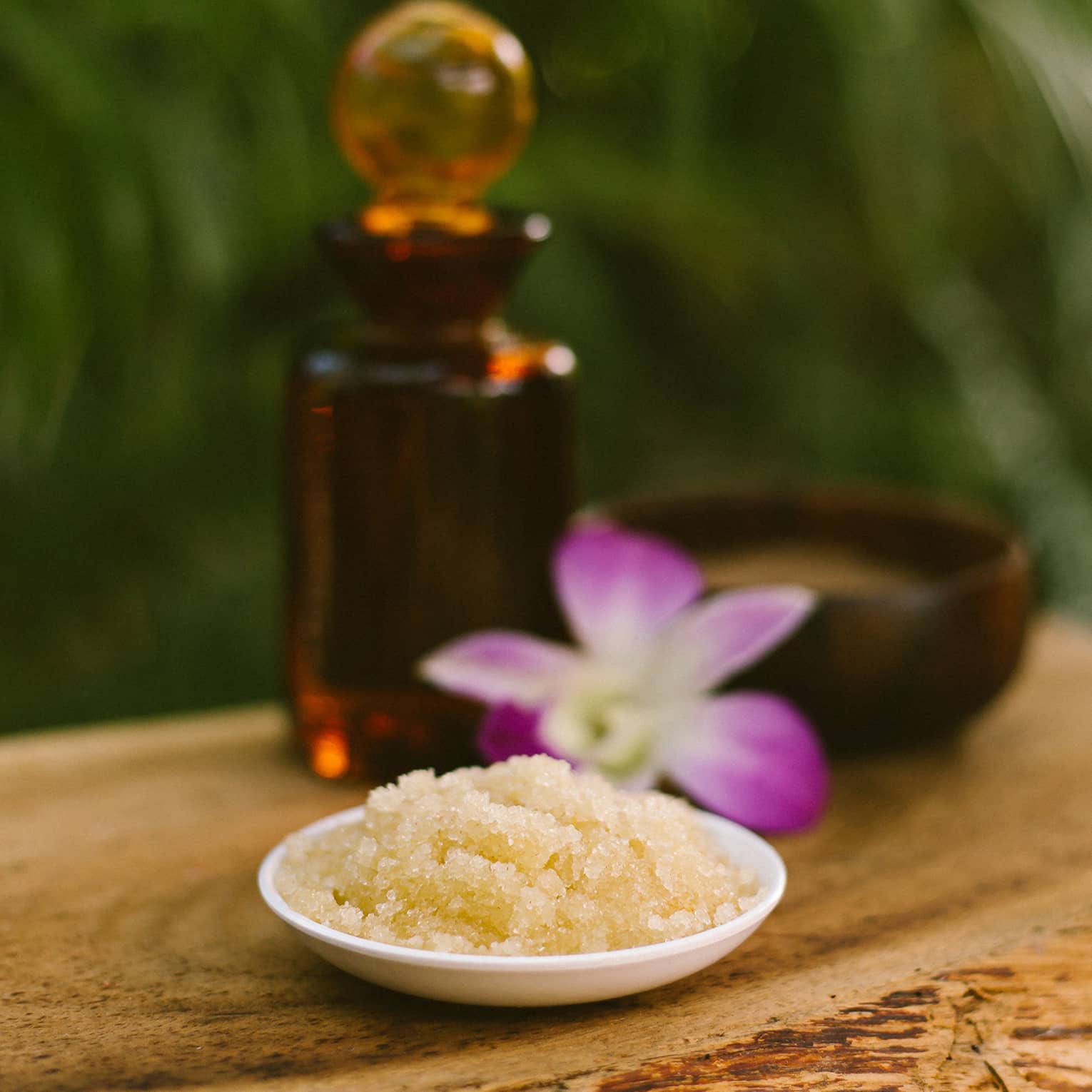 Bowl with salt scrub next to purple tropical flower and glass oil bottle on rustic table