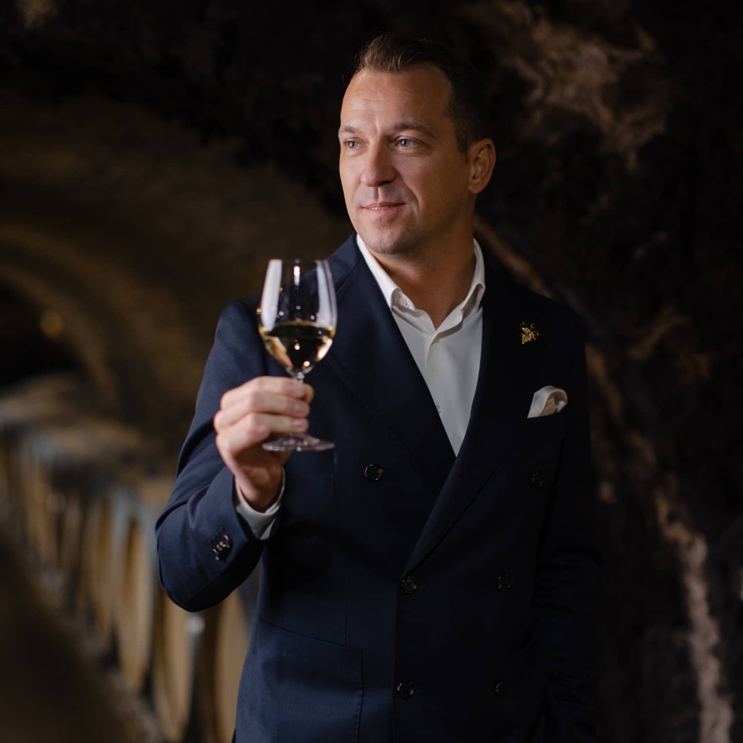 Person in a dark suit holding a glass of white wine in a wine cellar