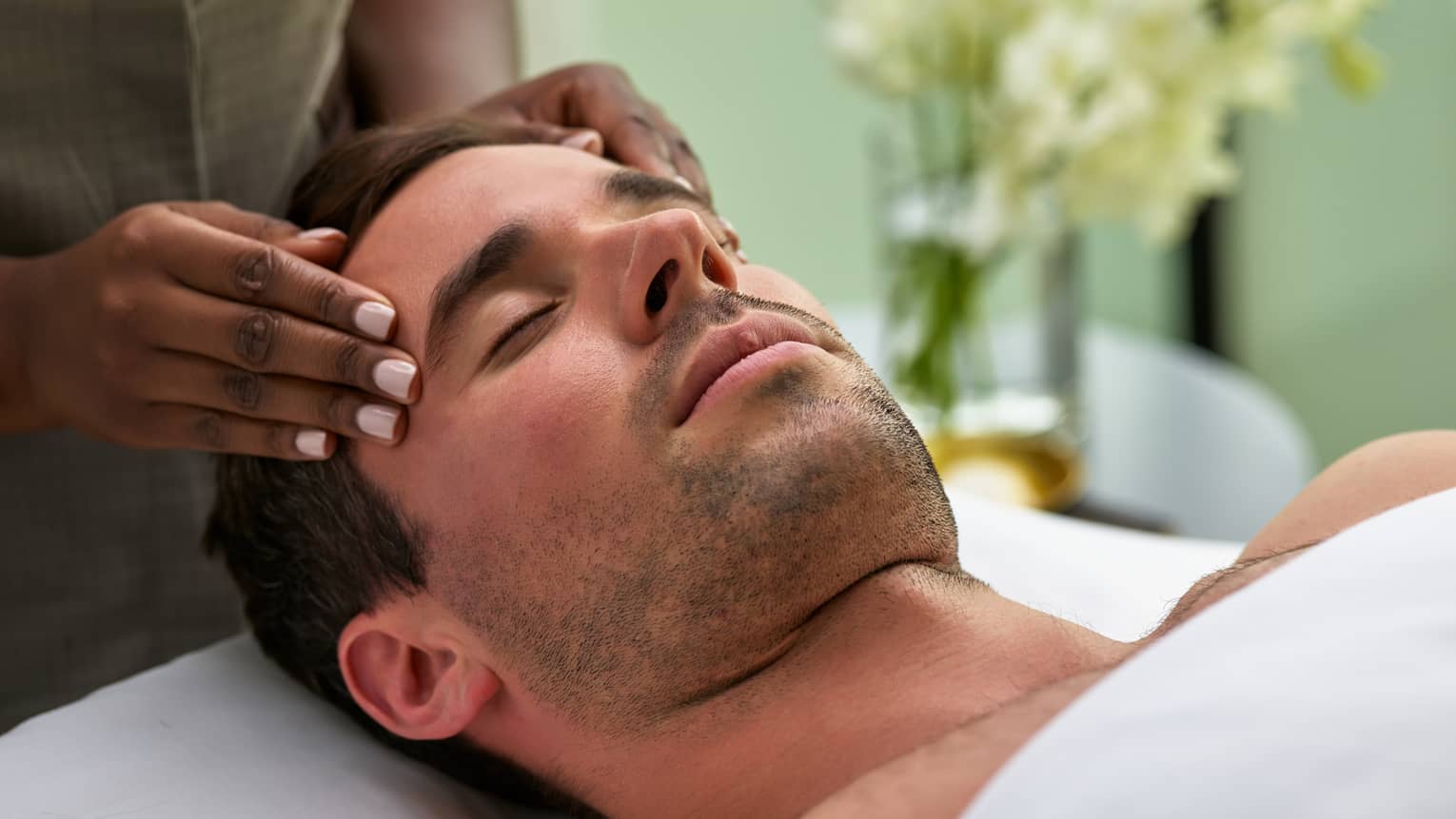 Man lies on massage table with eyes closed as hands massage his temples, forehead