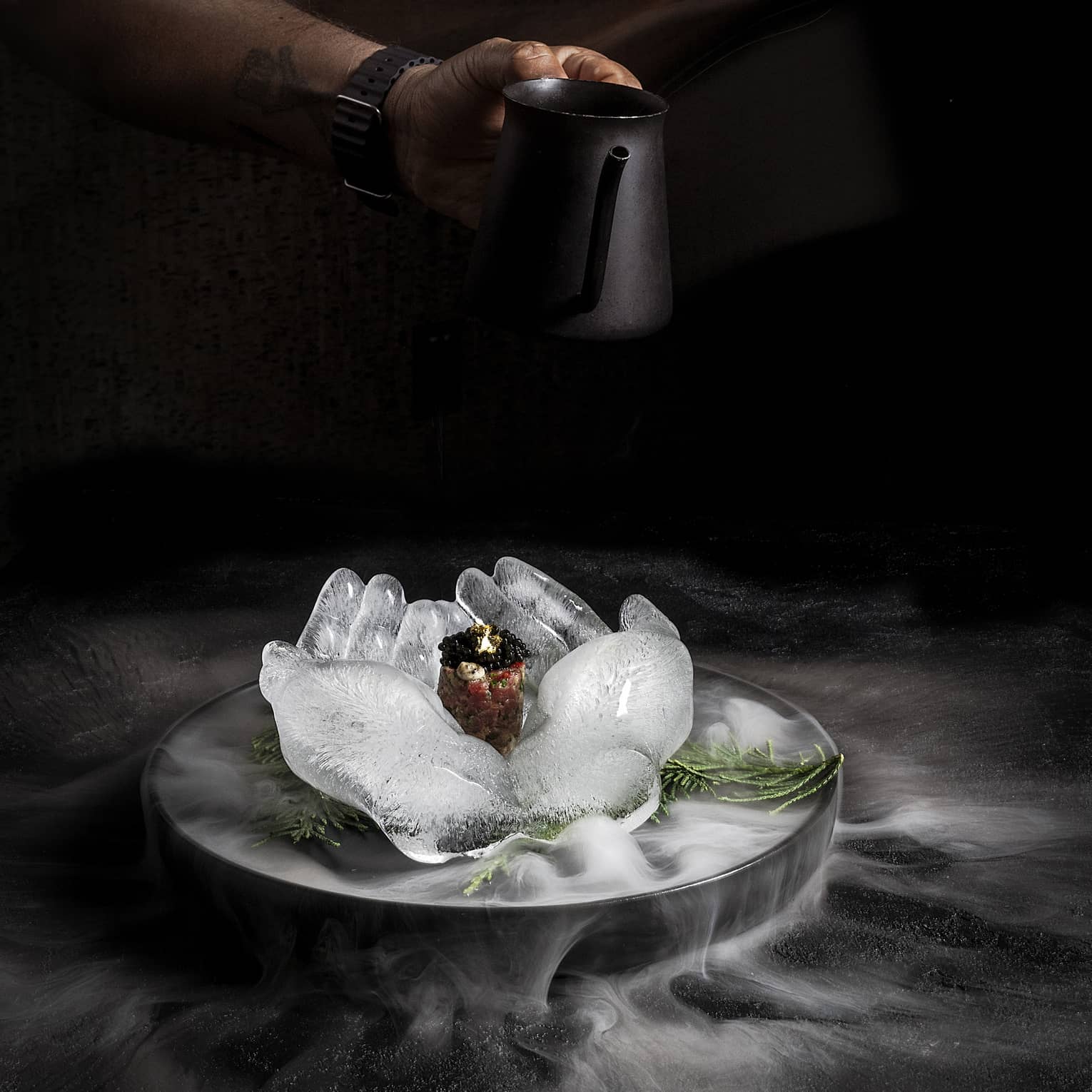 Food served on an ice sculpture shaped like two open hands with steam from dry ice floating around the black background