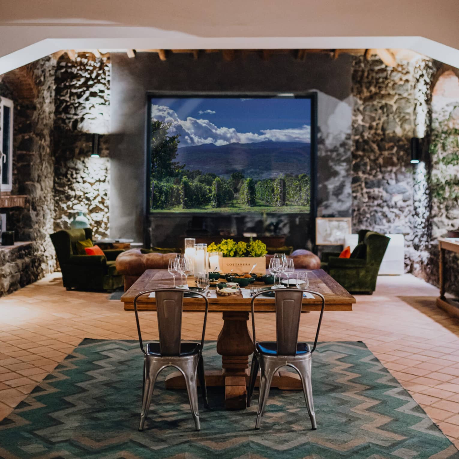 A wooden dining table set with floral arrangements and glassware, in a room with a window overlooking mountains and a vineyard