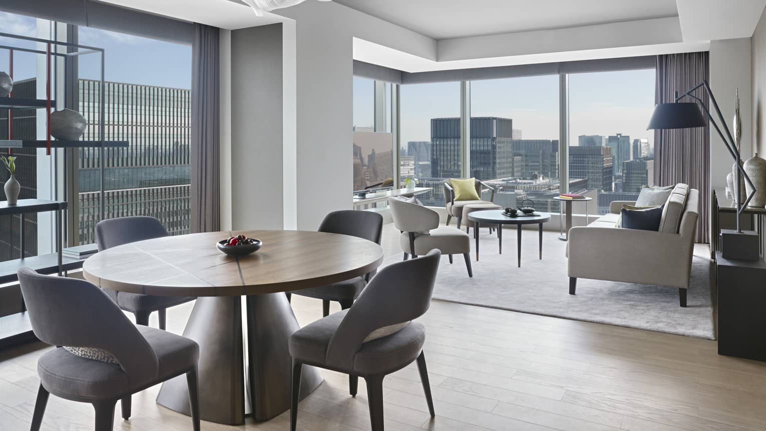 Hotel suite with round dining table with chairs, seating area, modern shelving and floor-to-ceiling windows with panoramic city views