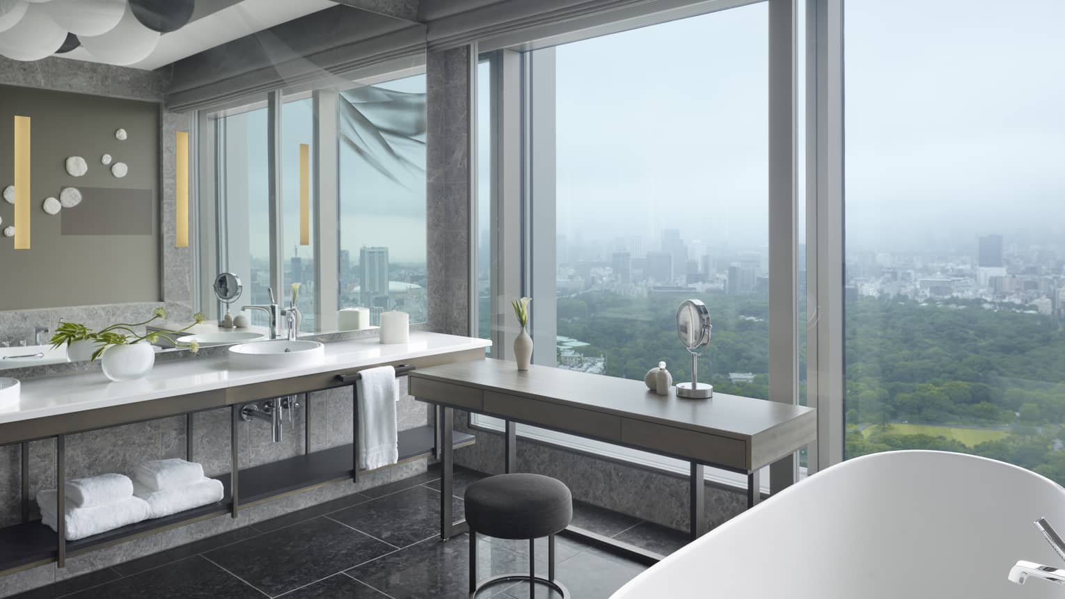 Luxurious bathroom with a double-sink vanity, bathtub, seating area and floor-to-ceiling windows offering expansive view of the city and lush greenery