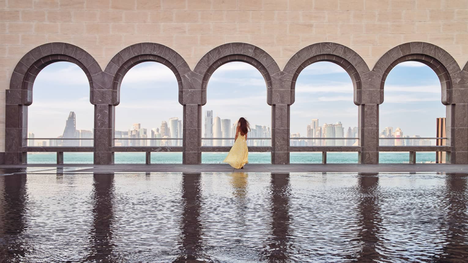 Woman wearing a long flowing yellow dress stands at the edge of a fountain beneath a wall of archways looking out onto the sea and a city skyline