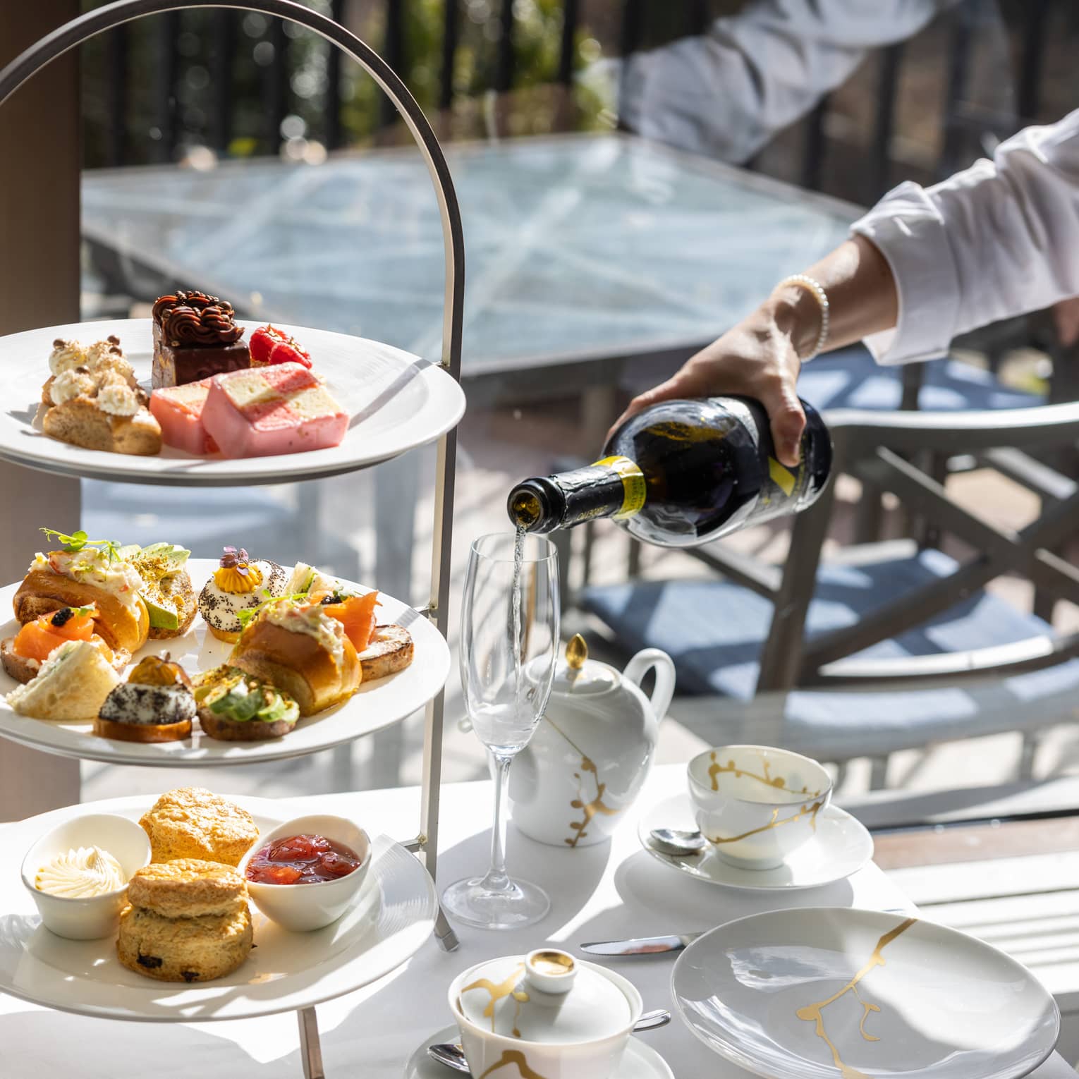 Champagne being poured next to a tiered assortment of afternoon tea small foods and pastries.