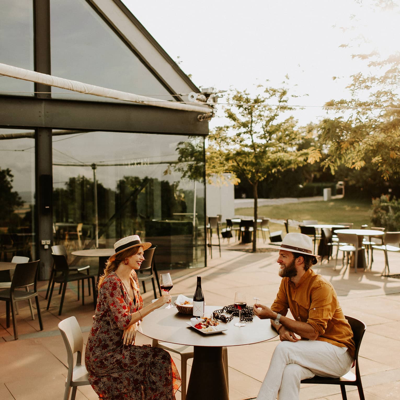 Couple wearing hats sit at outdoor table with plate of food, drinking red wine