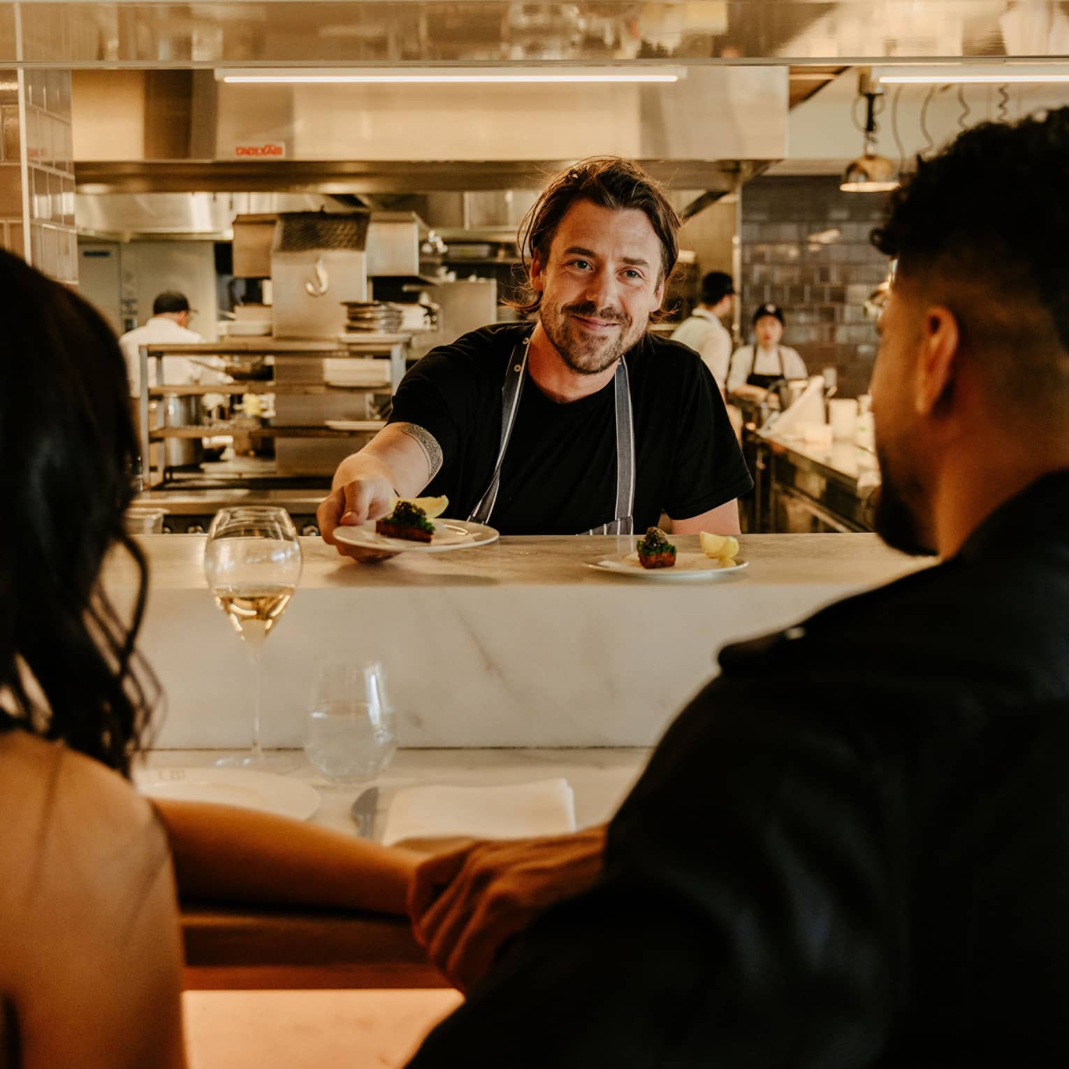 Rear view of a couple sitting at a counter as a smiling chef hands them a small plate, a blurred kitchen in the background.