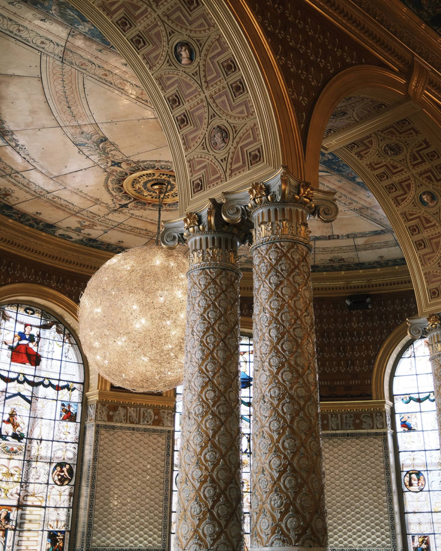 An interior space with a high arches and columns with decorative inlay and carving. Stained-glass windows, decorative ceiling and walls, and chandelier lighting are in the background.
