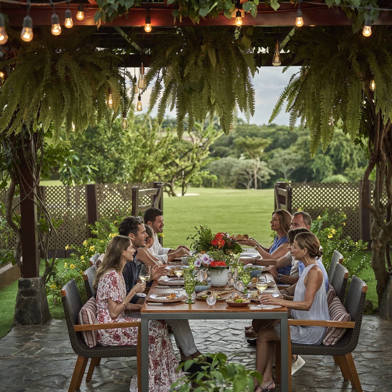 Eight people sit around a long dining table set up outside beneath a canopy of greenery and string lights