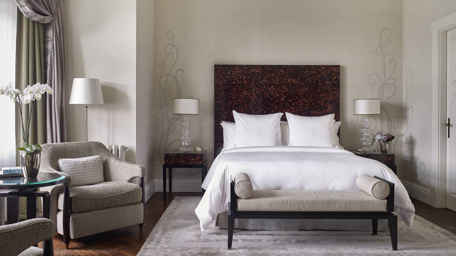 Elegant hotel bedroom with white bedding, dark wood headboard, and cozy sitting area with armchair and orchid on side table