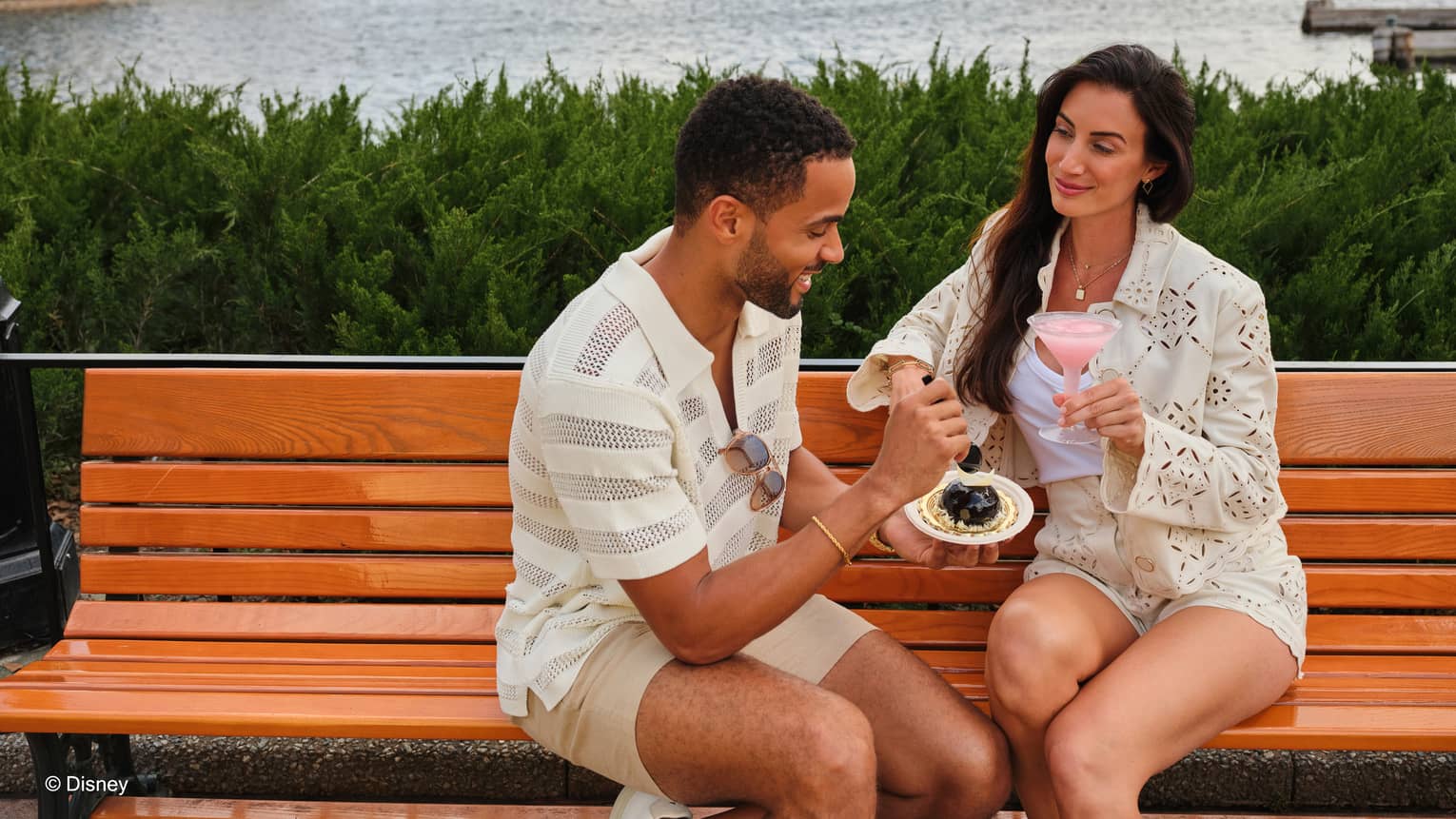 Couple sits on a park bench by the water sharing a drink and dessert