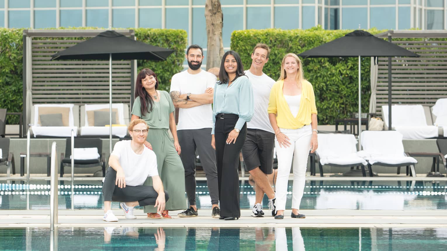 A group of six people are posing by the outdoor pool with a scenic view of a harbour in the background. They are dressed casually.