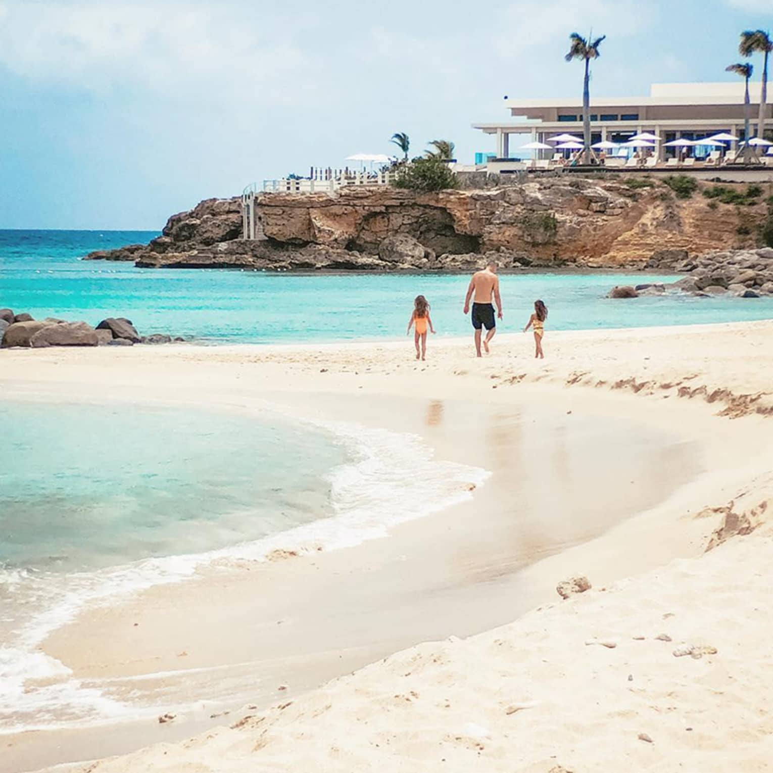 Person walking with two children along a sandy beach with clear turquoise water, rocky cliffs and a luxury resort building in the background
