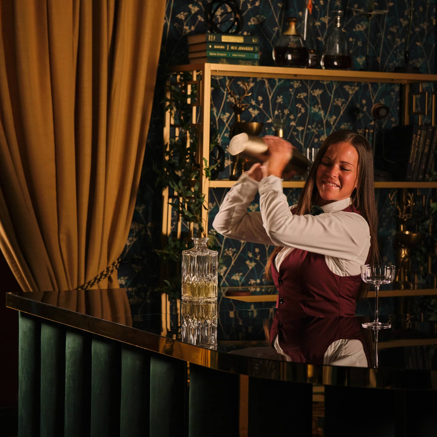 Bartender wearing white shirt, burgundy vest and black pants stand behind the bar shaking a cocktail
