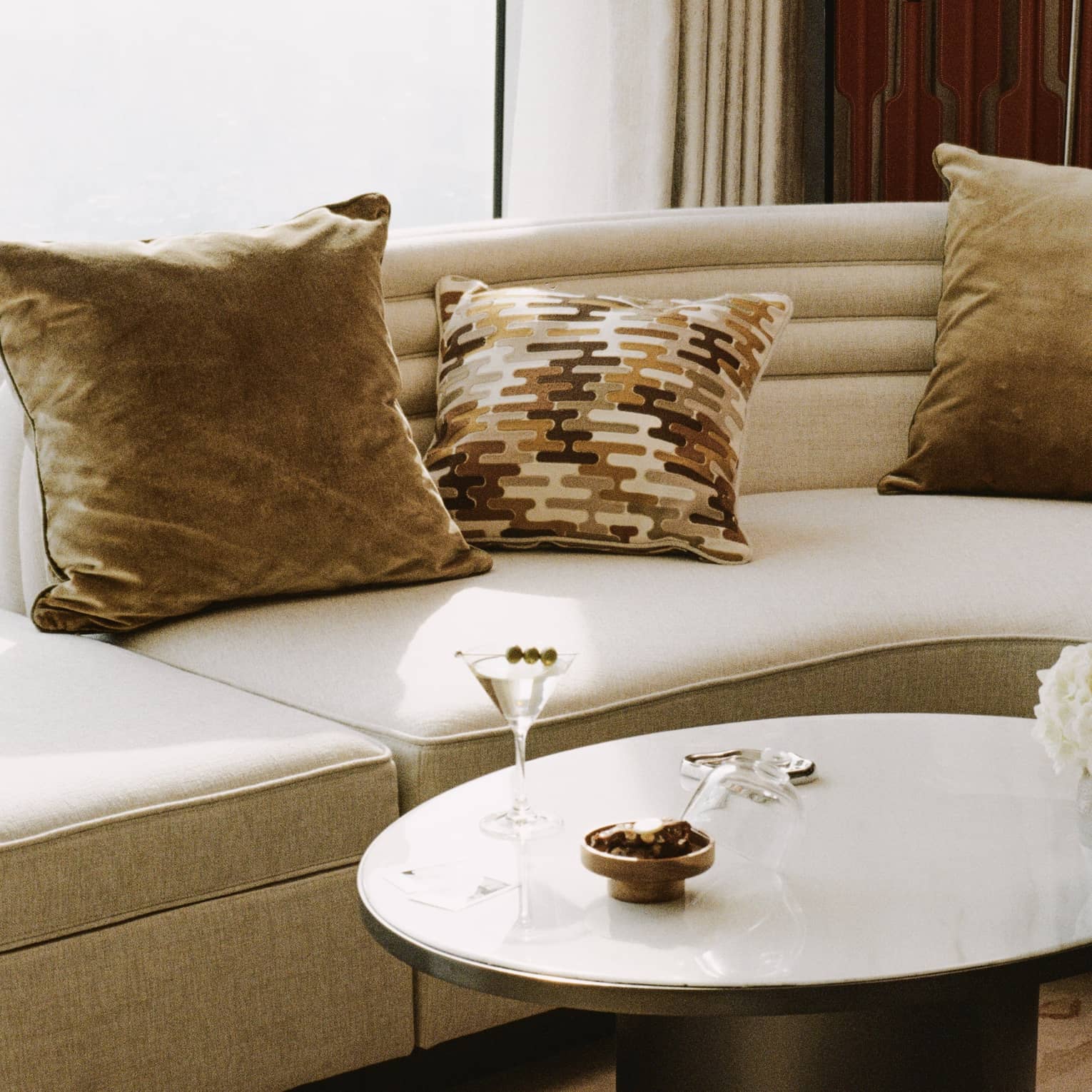 A corner of a room with a tan linen couch with brown accent pillows, a modern, circular coffee table topped with a martini and a bowl of nuts and an abstract printed rug