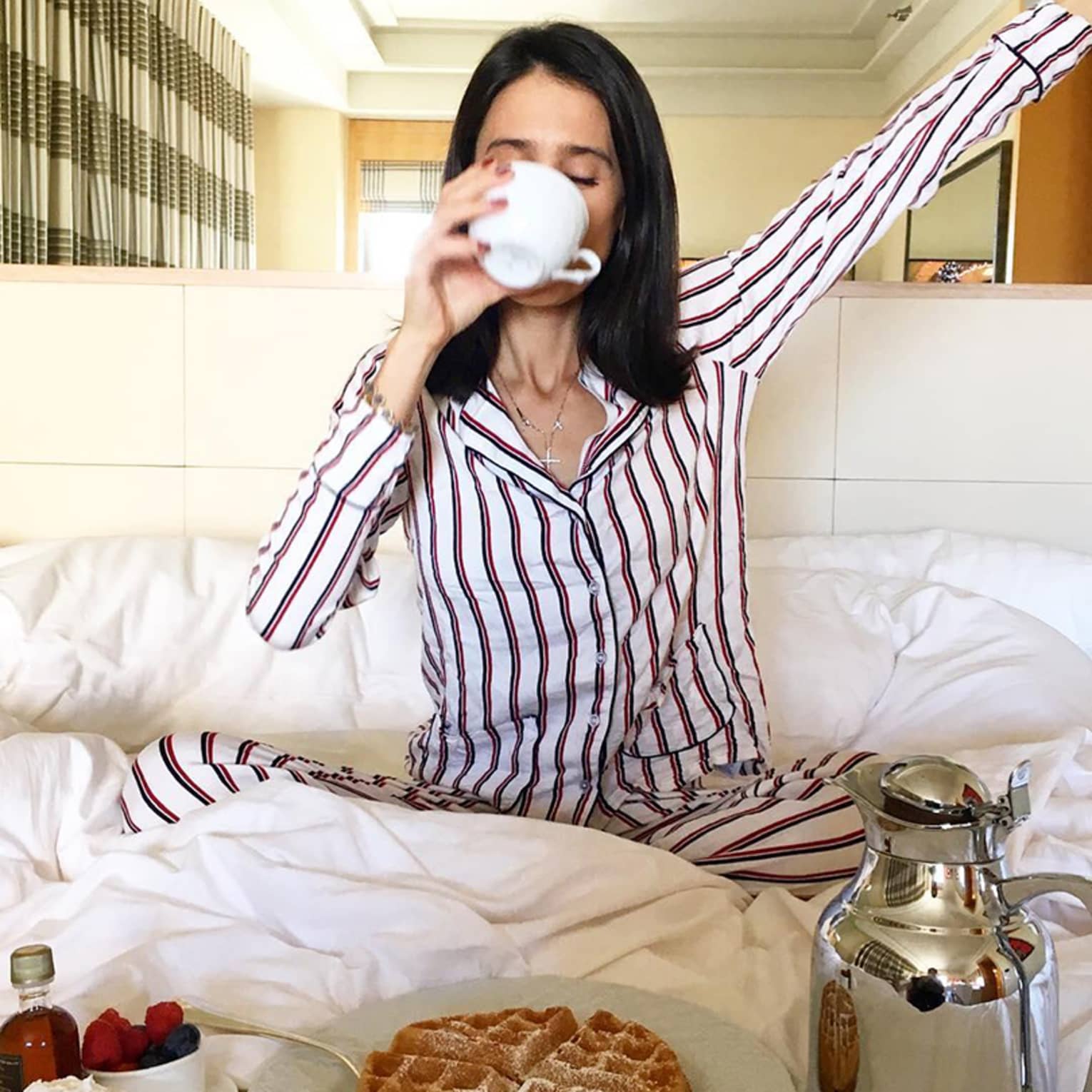 Woman on bed sips coffee cup, stretches in front of in-room breakfast dining tray