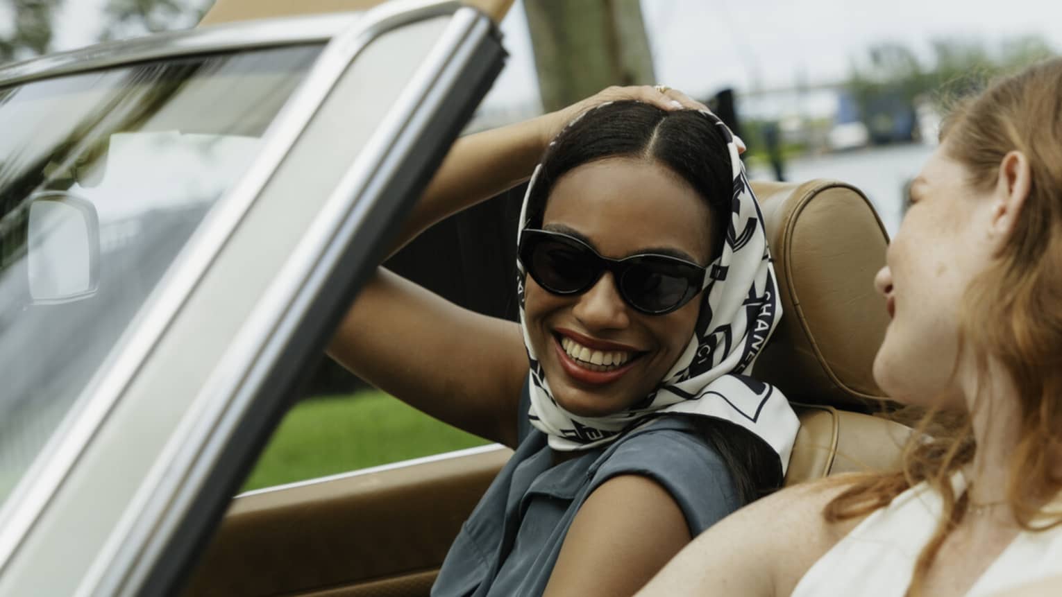 Two guests riding in a convertible car while smiling at one another.