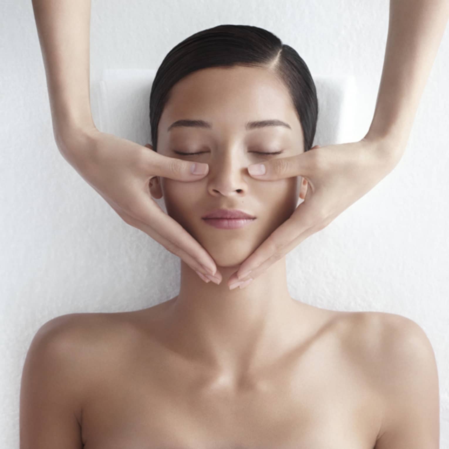 Woman in white bathrobe lies on massage table with eyes closed, masseuse rests hands on her cheeks