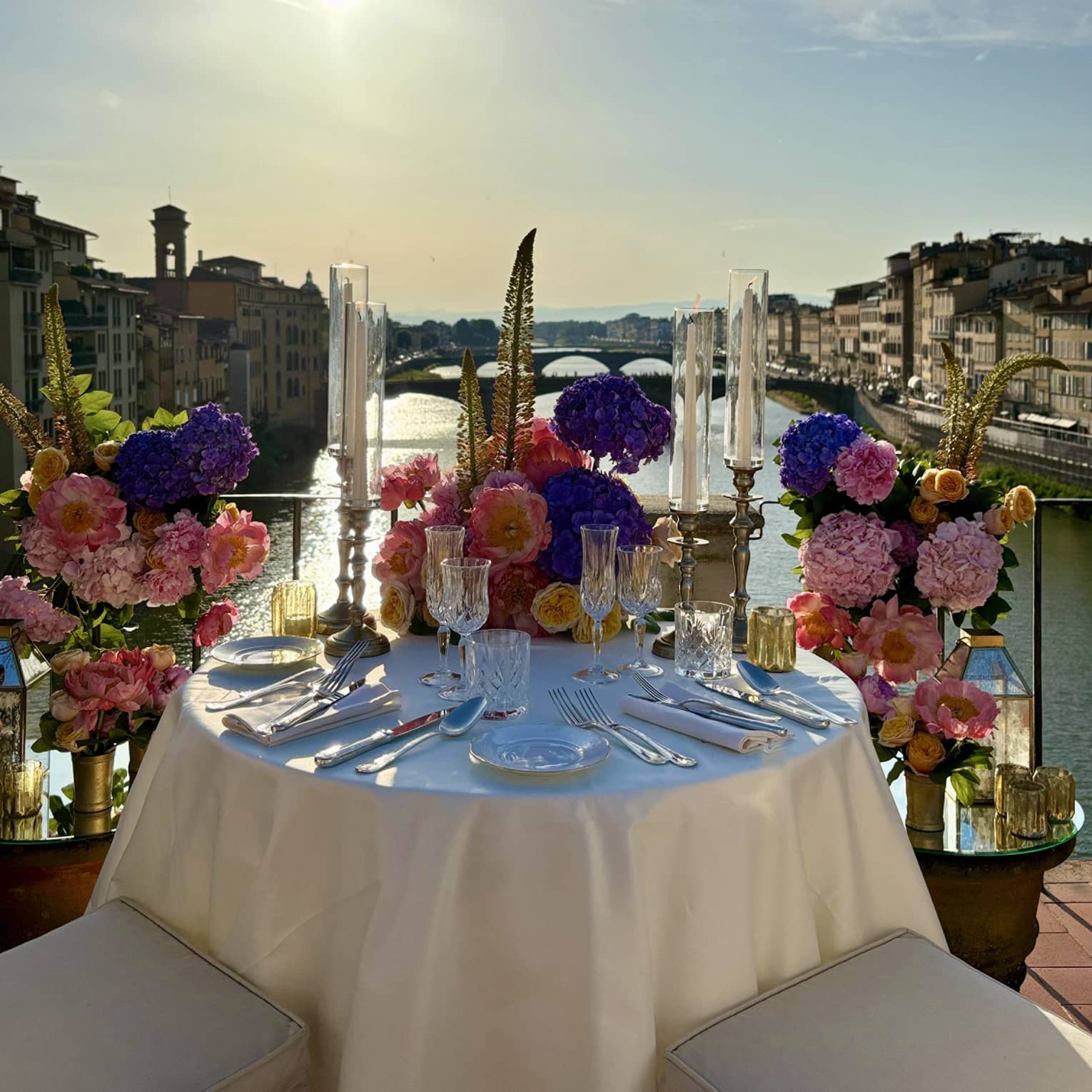 An elegantly set outdoor dining table overlooking a a river, with a sunny sky in the background.