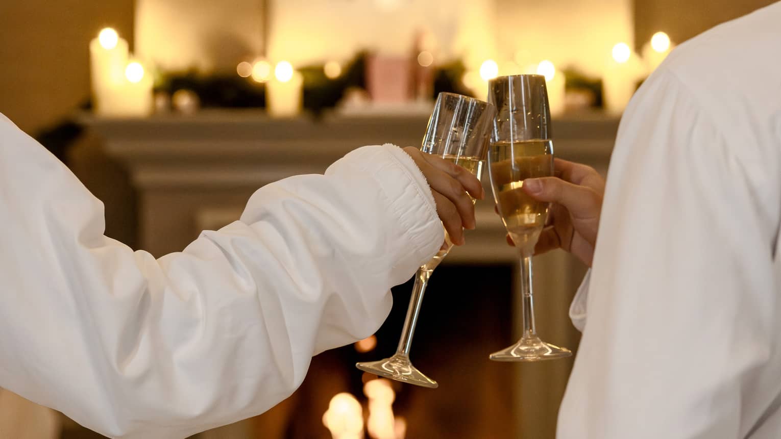Two guests wearing white robes sit in front of a fireplace toasting each other with glasses of Champagne