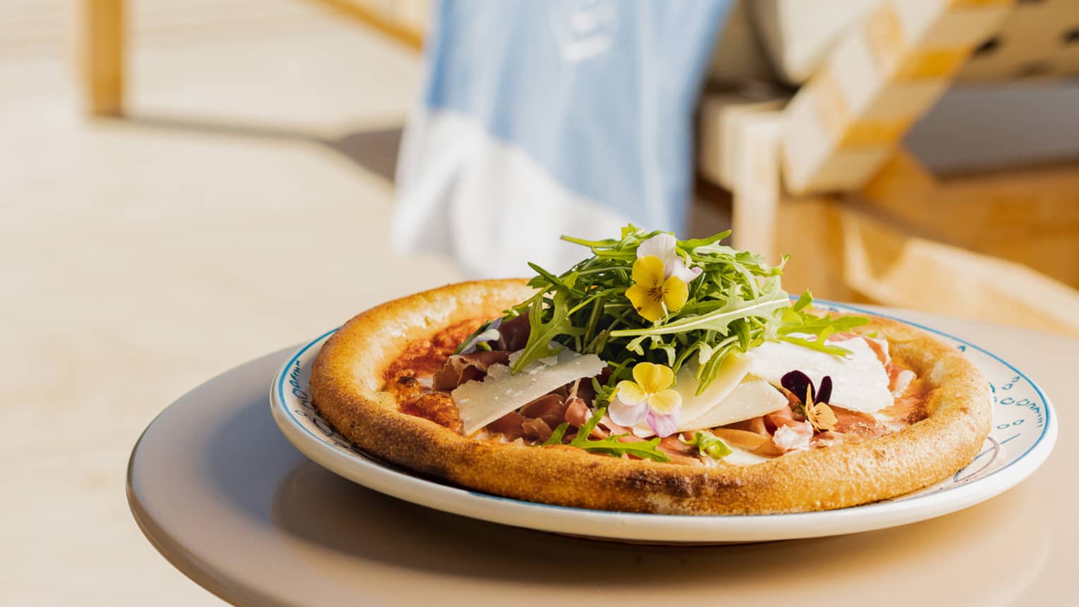 Personal-size pizza topped with microgreen garnish on round outdoor table near lounge chair by pool