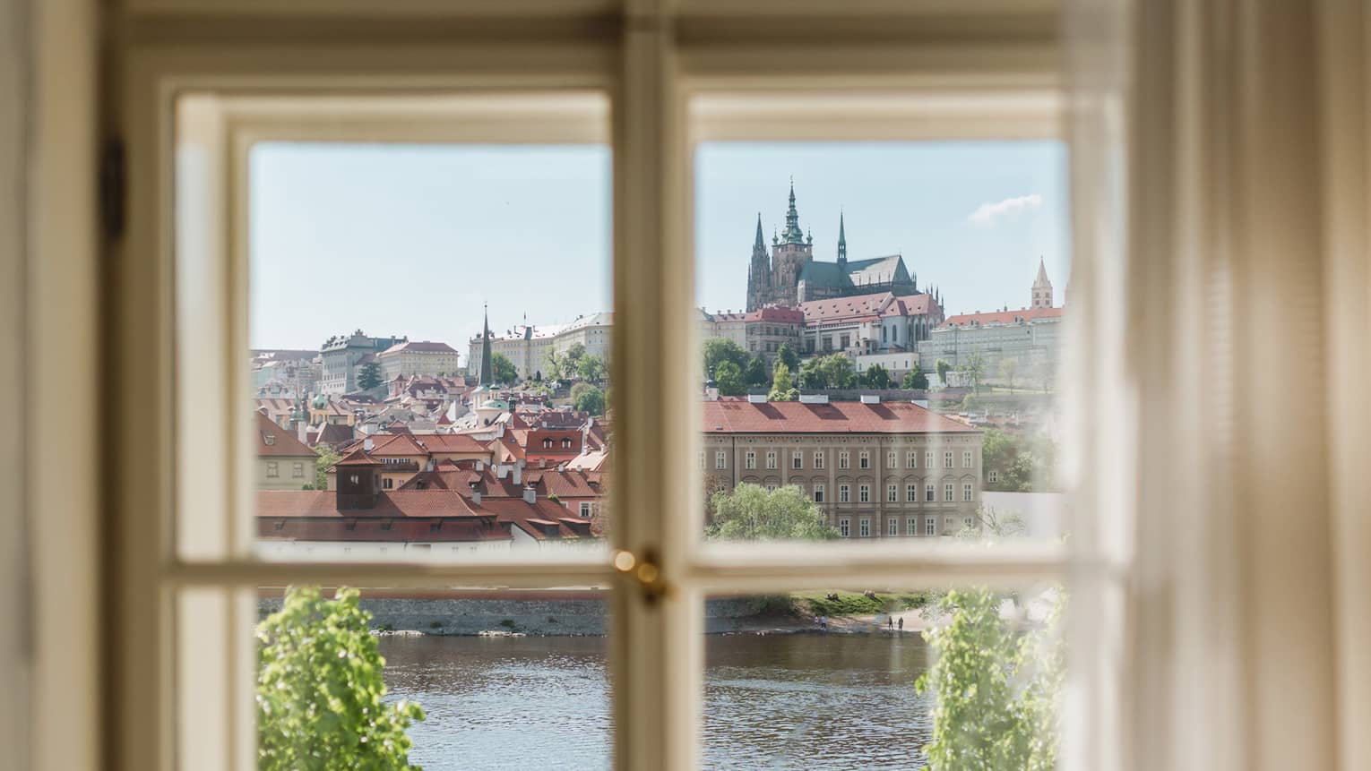 View through a window of a historic city with red rooftops and a cathedral in the distance, overlooking a river and green trees under a clear blue sky.