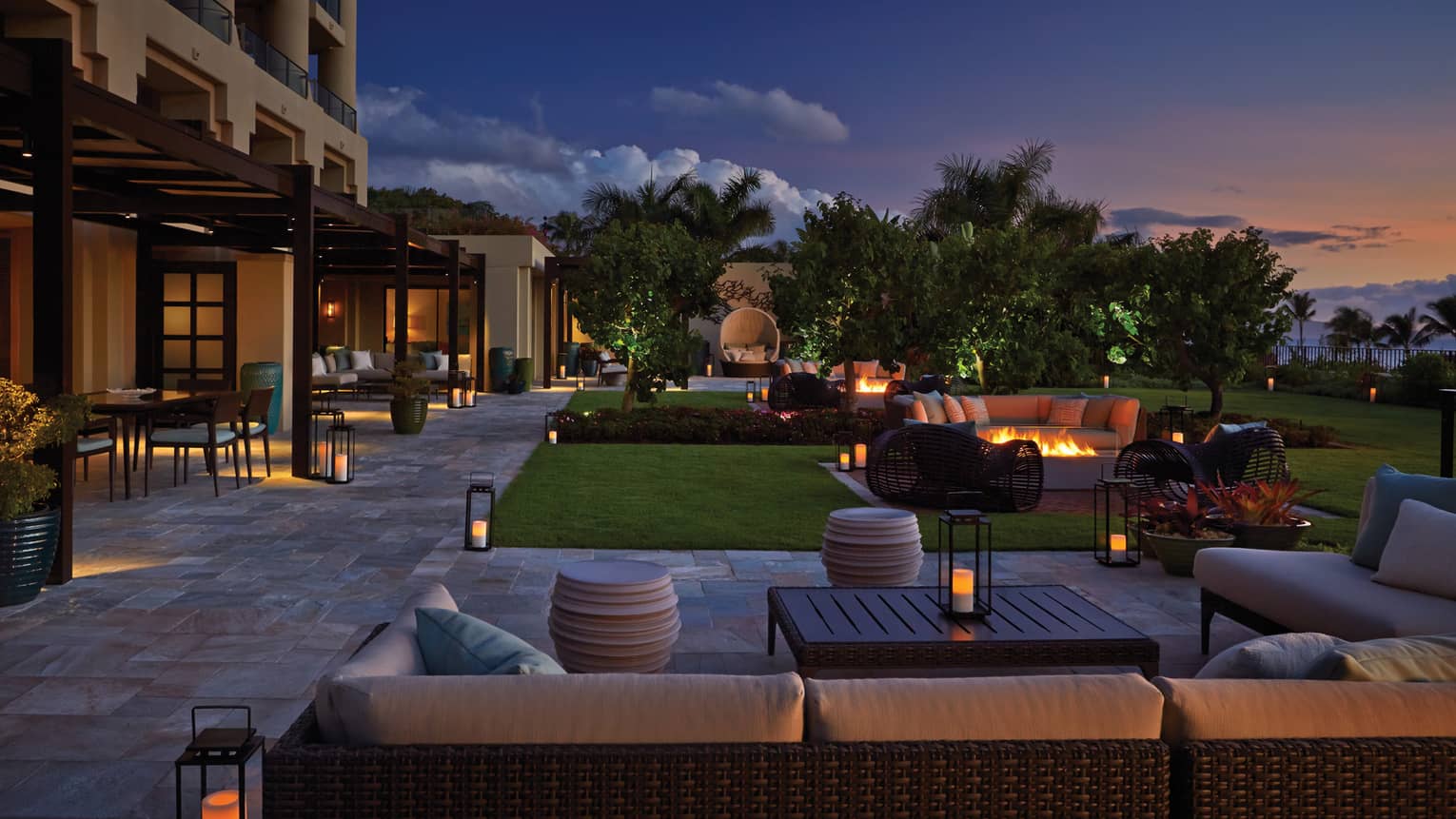 Outdoor lounge area at dusk with wicker seating, fire pits, lanterns and garden landscaping