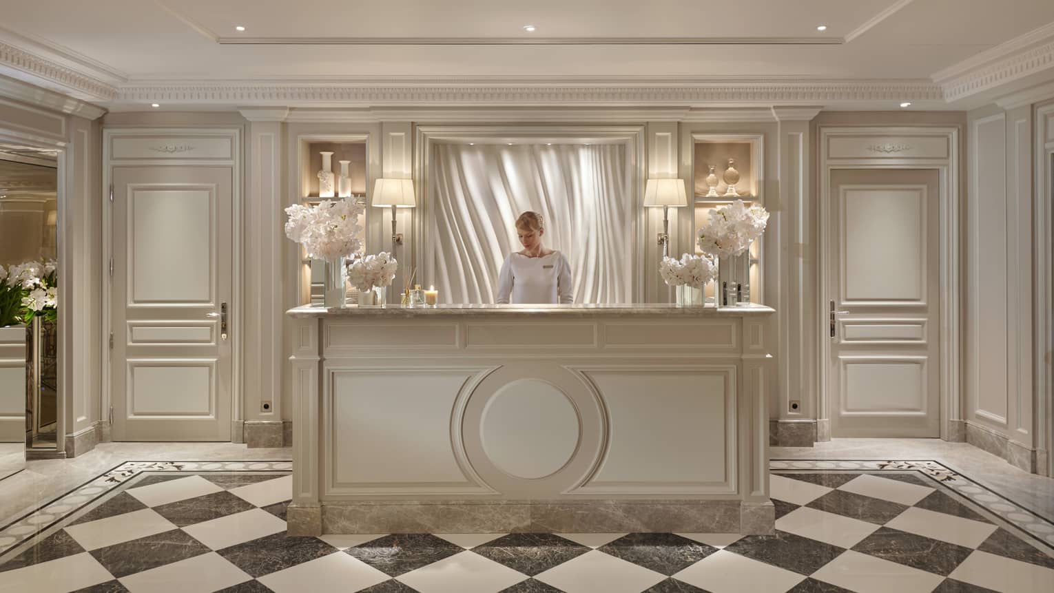 Receptionist standing behind spa reception desk atop checkerboard marble floor amid ambient lighting