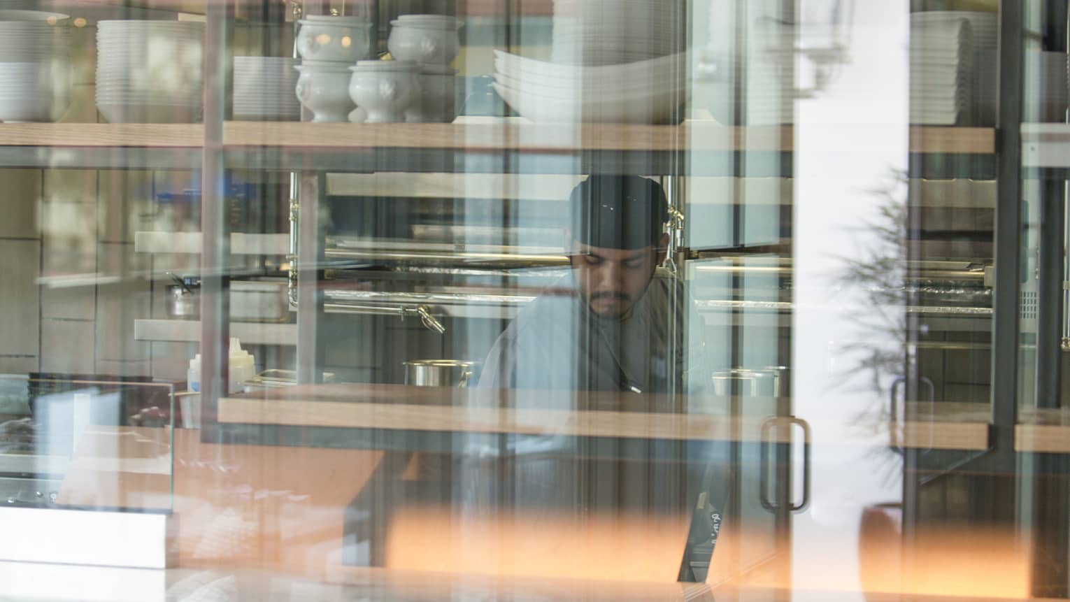 A chef working in a kitchen behind large glass windows, there are stacks of white dishes behind him.