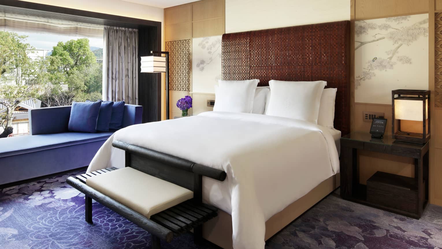 Elegant bedroom with a king bed, a purple chaise lounge, rug and flowers and floor-to-ceiling windows overlooking lush greenery