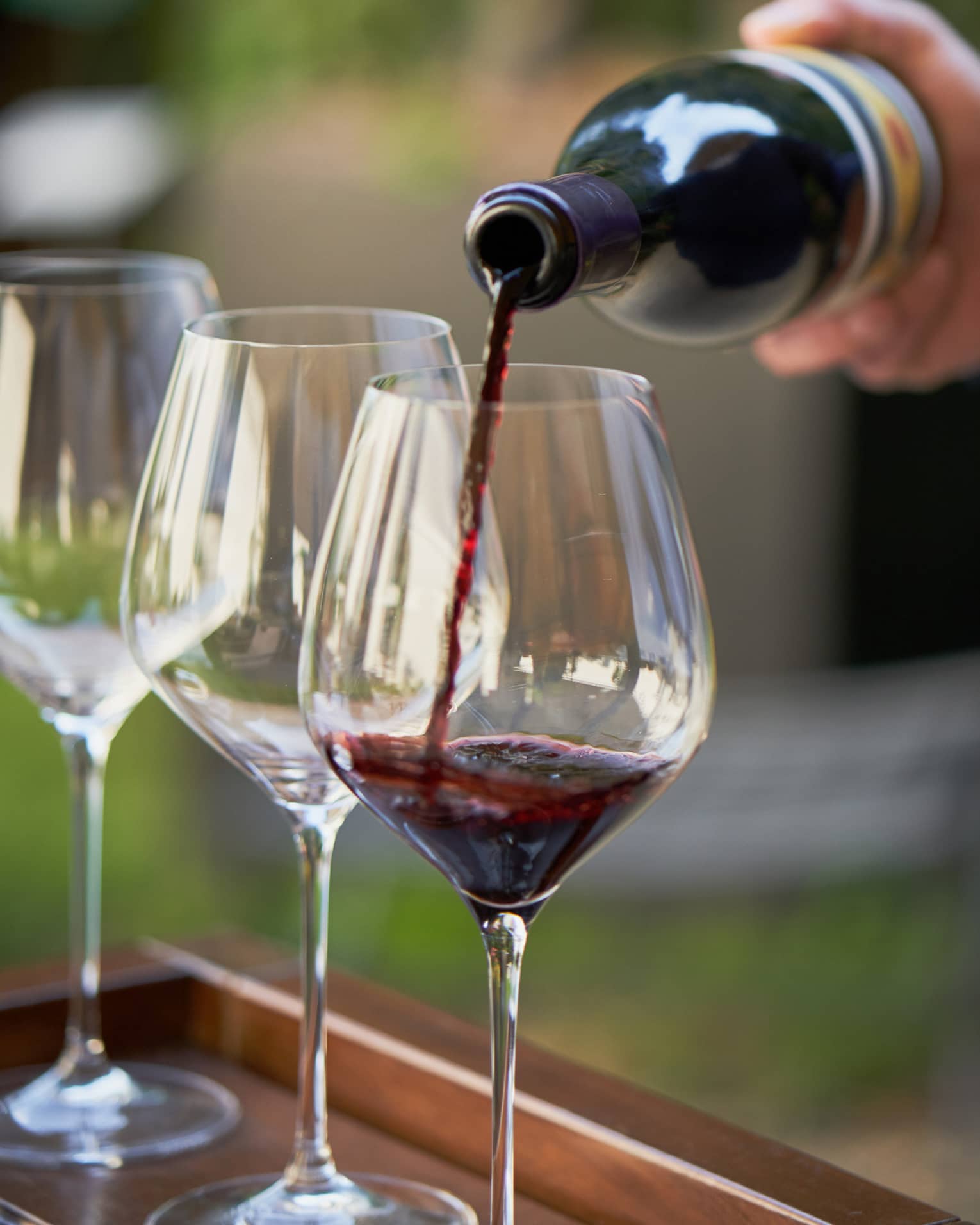 A close up of hands pouring a bottle of red wine into a clear wine glass that rests on a wooden tray, outdoors