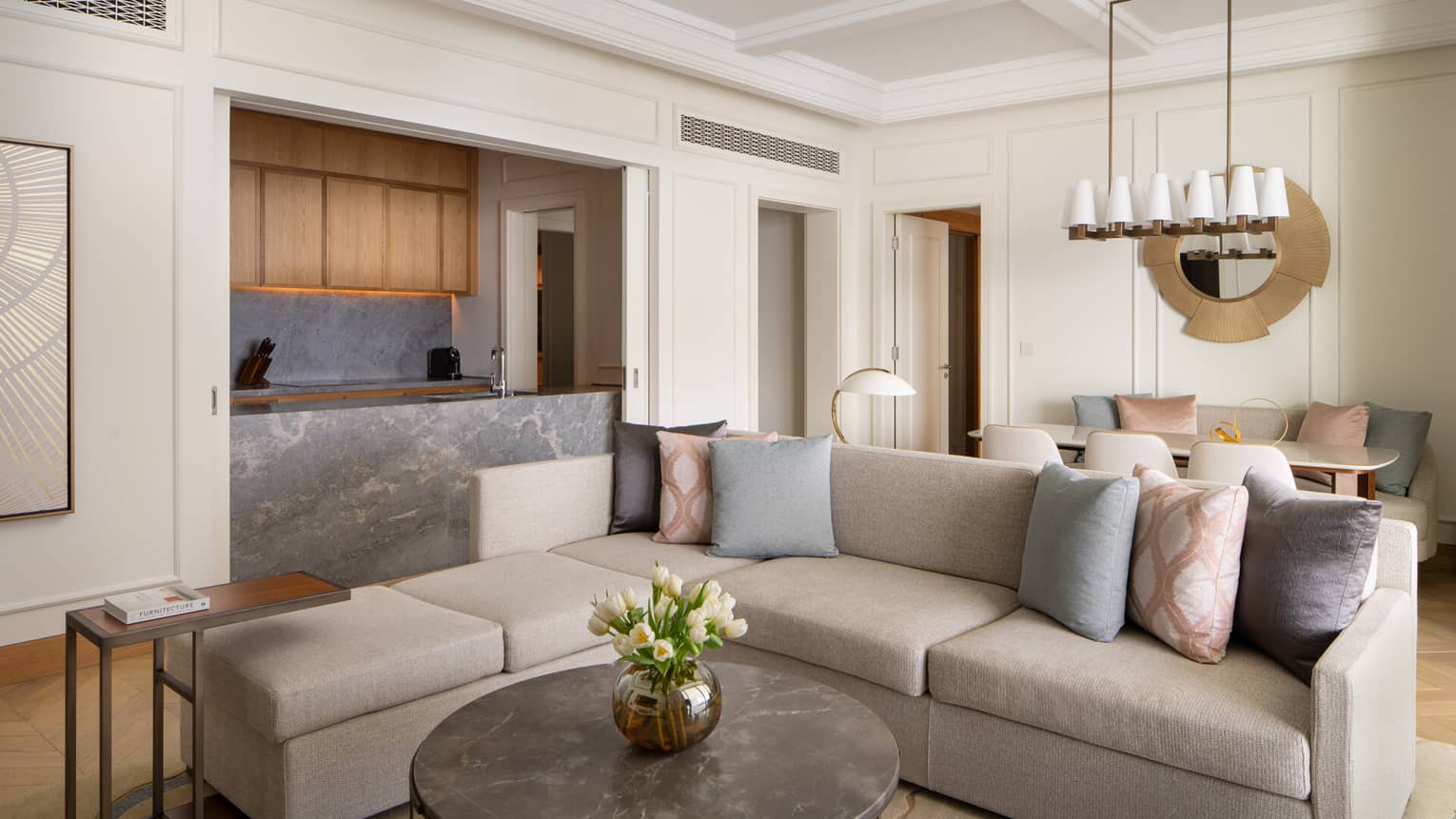 Contemporary living room with a grey sectional sofa, marble coffee table, open kitchen with wood cabinets and a dining area with pastel cushions.