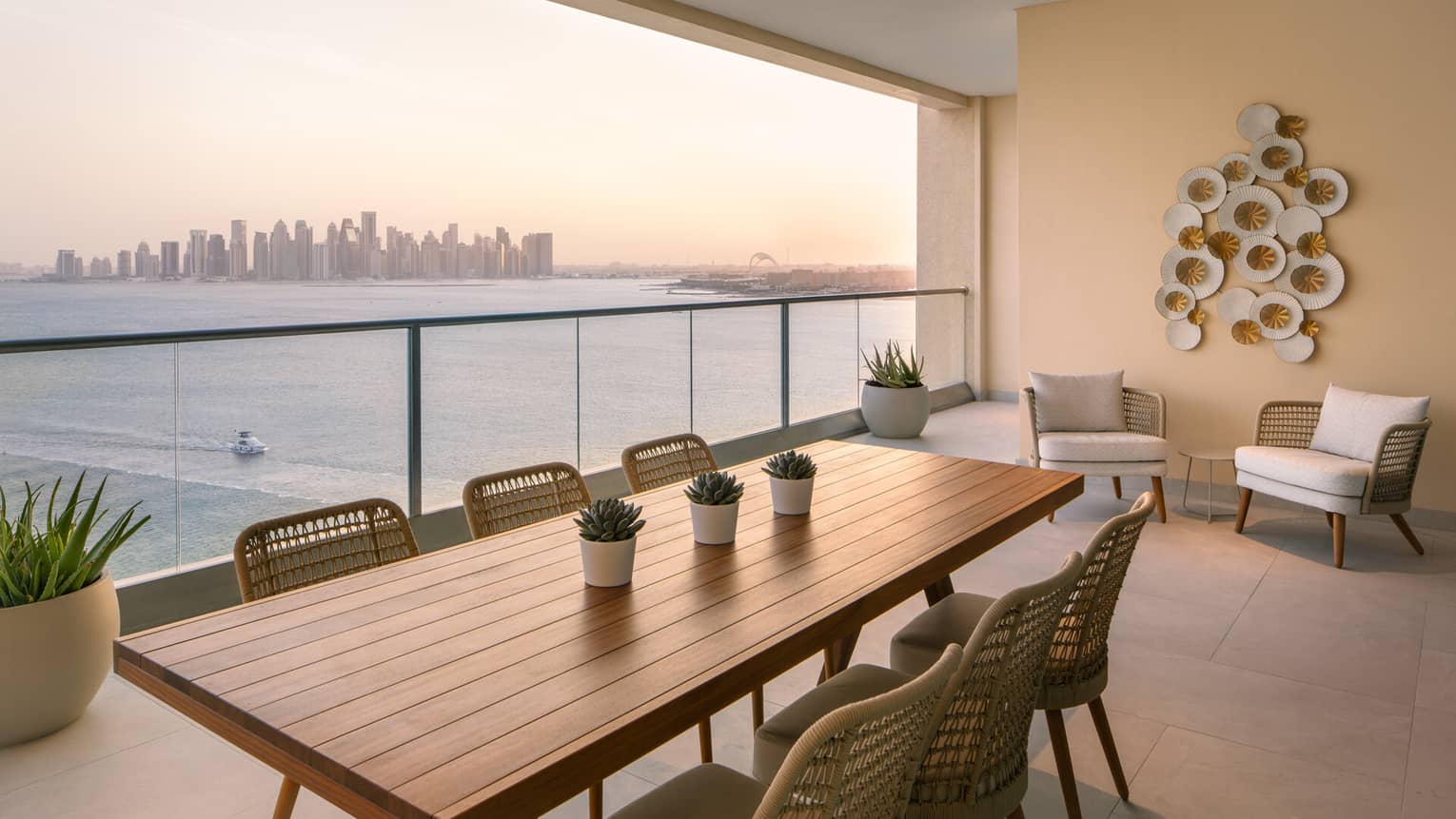 Modern balcony with woven chairs, a wooden dining table, potted plants and a sea view at sunset overlooking a city skyline.