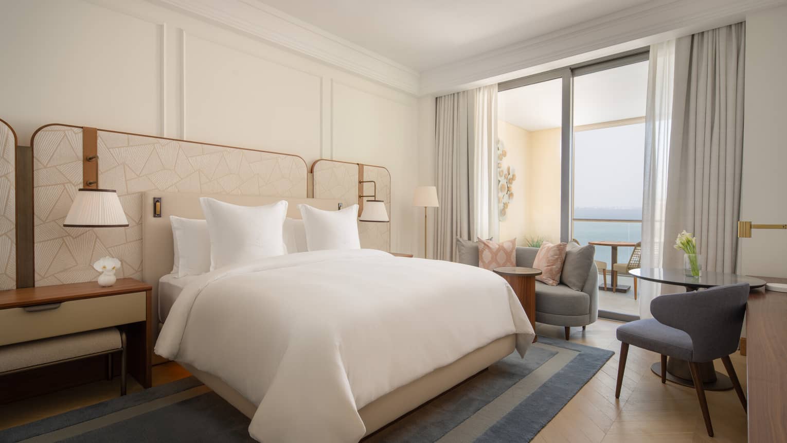 Bright bedroom with a large bed, neutral tones, balcony seating area and sea view through sliding glass doors.