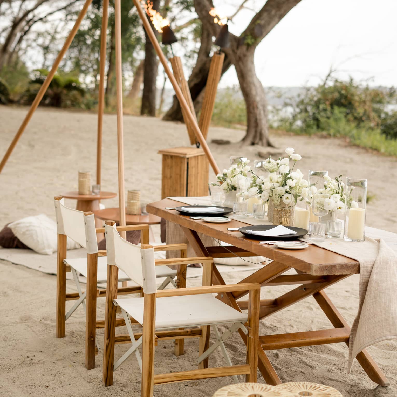 Wooden dining table and chairs set up on a beach, decorated with white floral centrepieces and candles