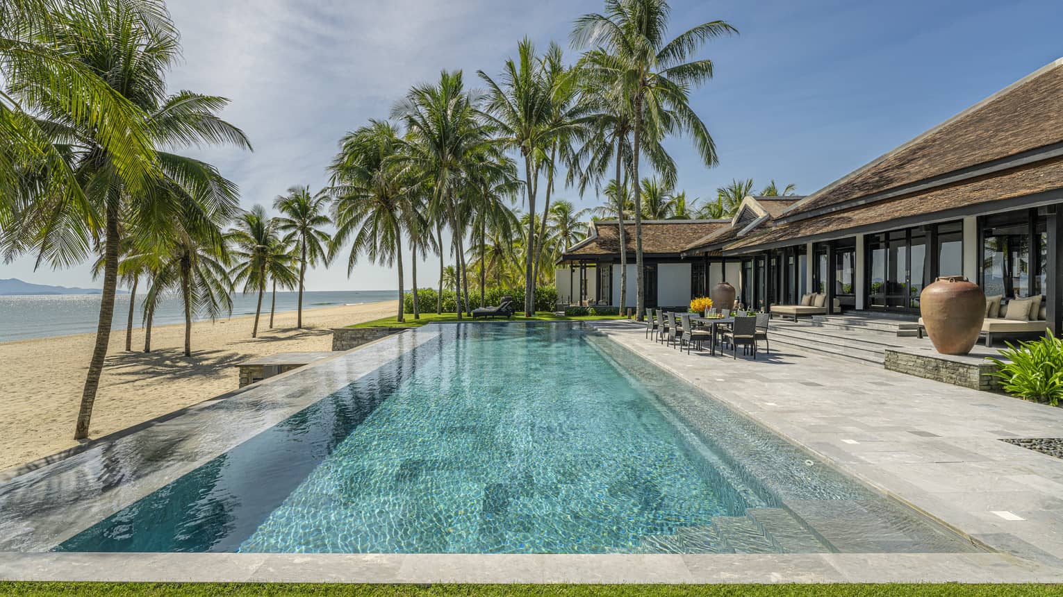 Luxury beachfront villa with a spacious pool surrounded by palm trees, overlooking a sandy beach and calm ocean