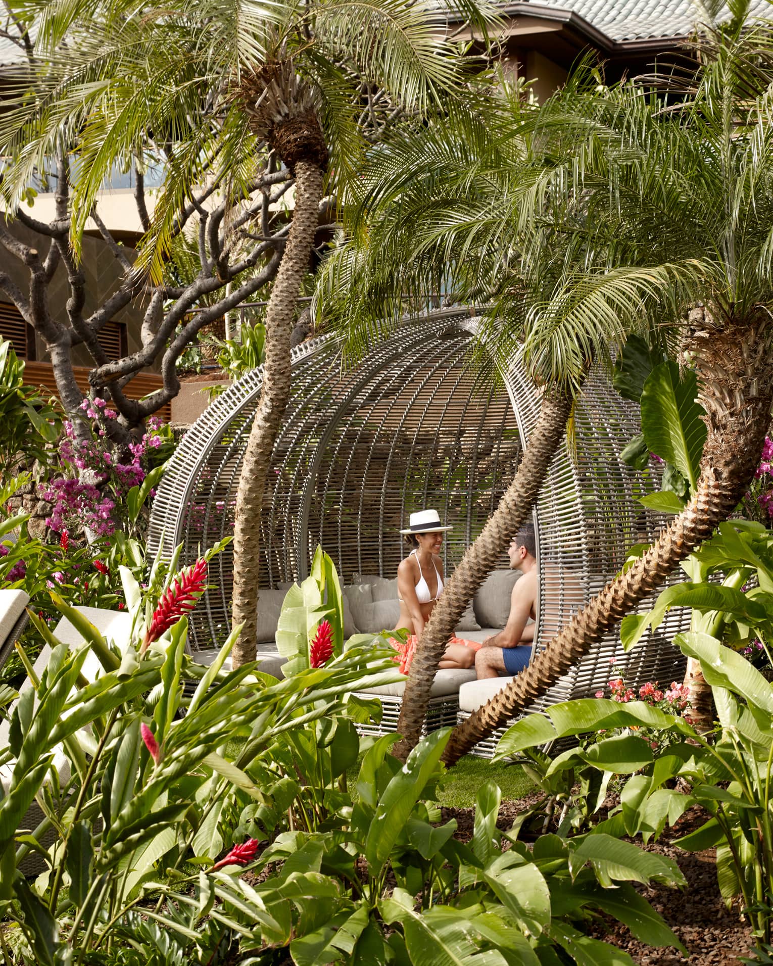 Couple sits in large wicker cabana in Pool Sanctuary outdoor patio lounge, tropical gardens