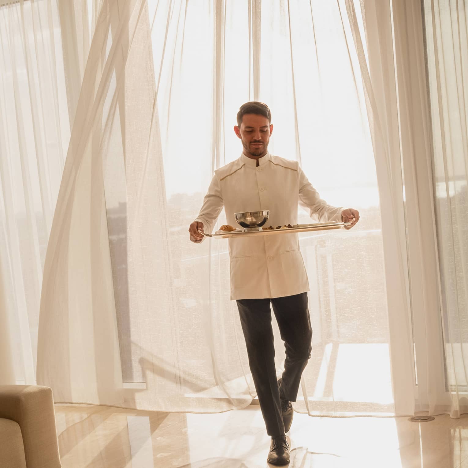 Hotel staff member carrying a tray with food for in-room dining, standing in front of sheer curtains with sunlight streaming through