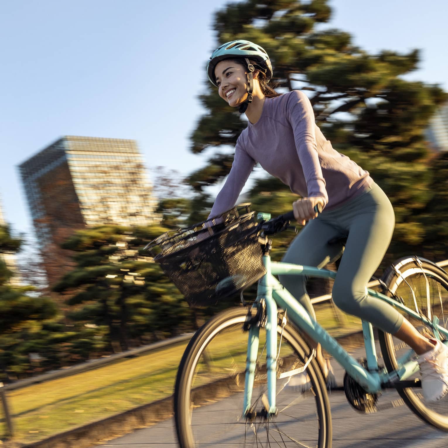 Smiling woman on teal cruiser bike with matching helmet rides by mature trees and skyscrapers of downtown Tokyo