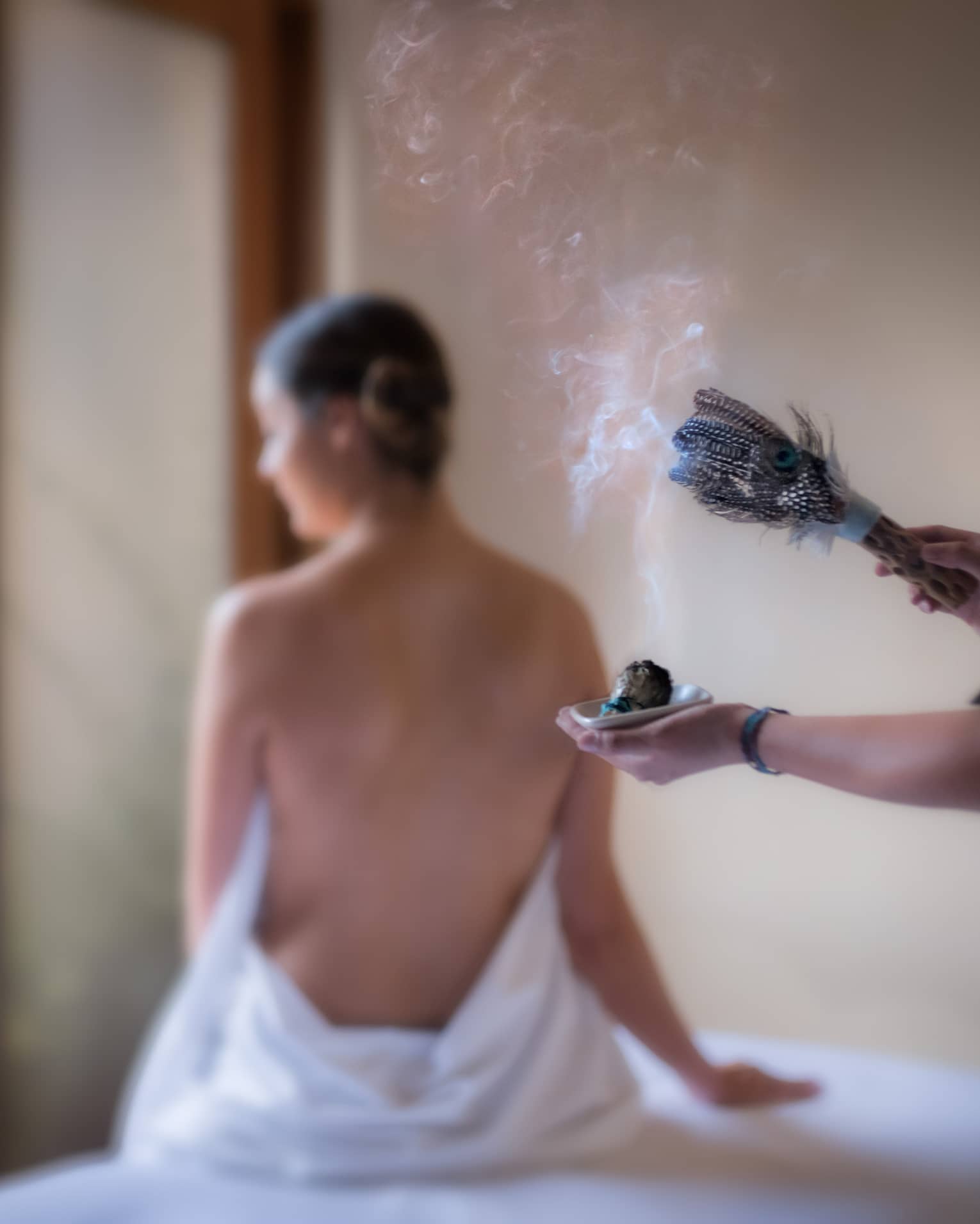 Smoke rises from sage brush with feathers as spa staff performs smudging ceremony for woman
