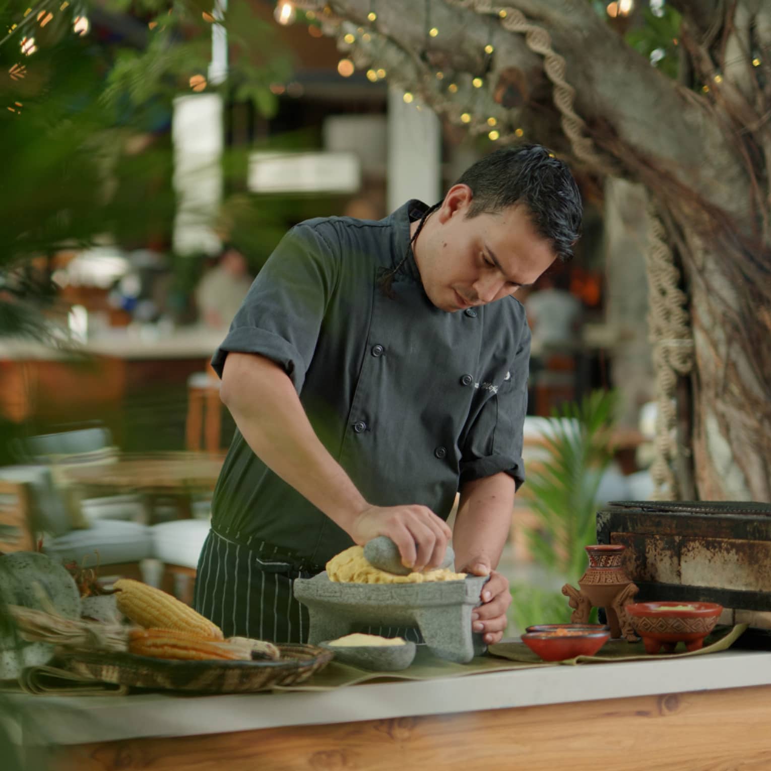 Chef wearing dark grey uniform mashes corn using a stone mortar and pestle