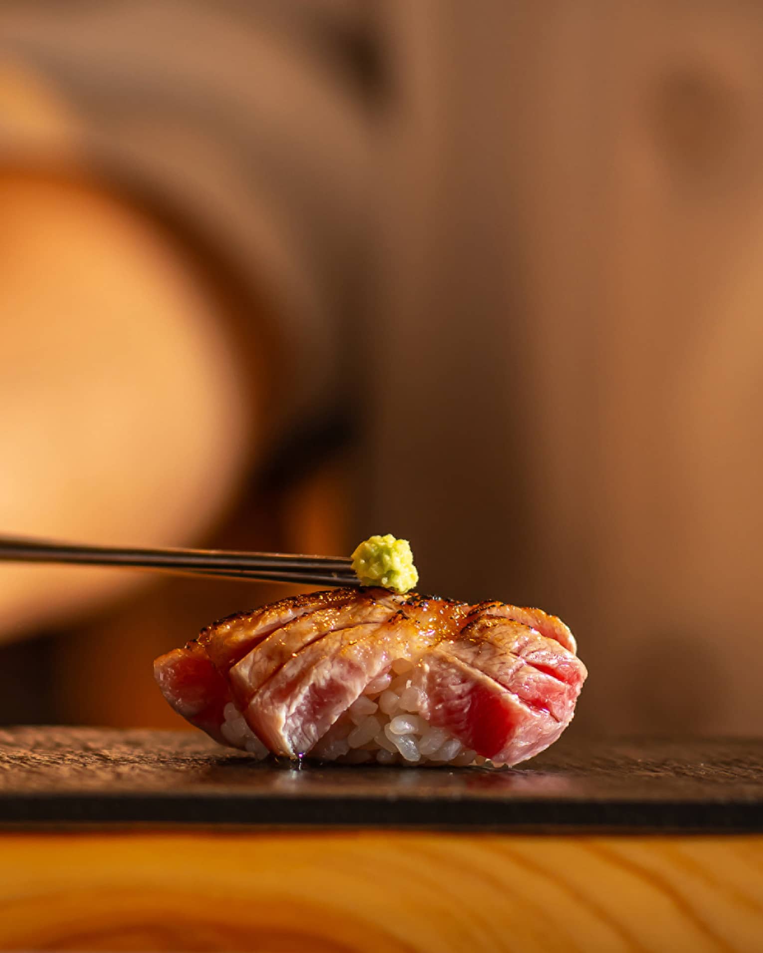 Seared tuna nigiri topped with a small dab of wasabi, placed on a black slate plate with a warm, blurred background
